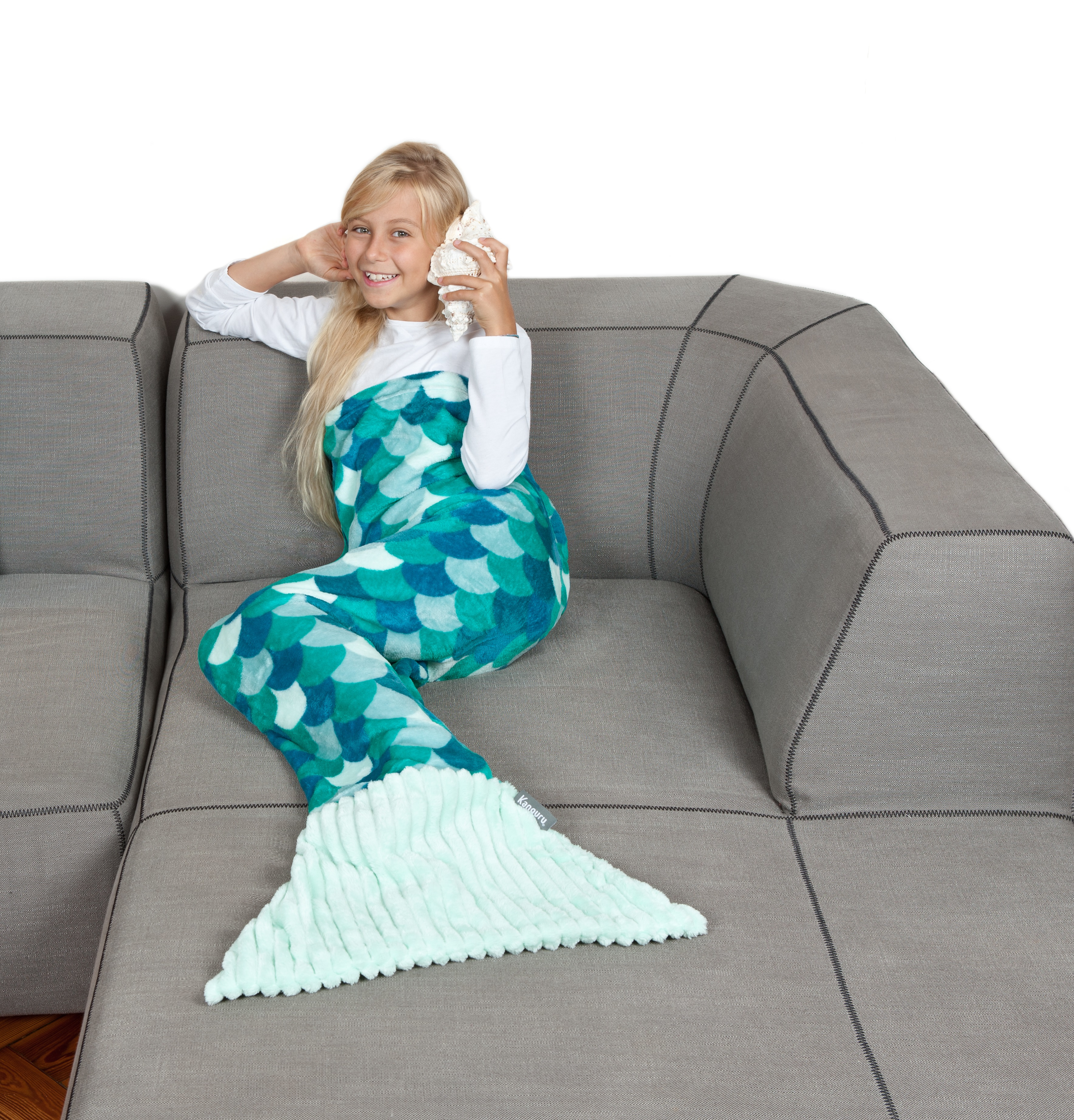 Una bambina in costume da sirena siede su un divano grigio, con una conchiglia all'orecchio.