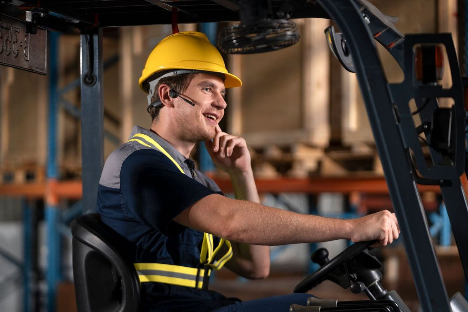 Mężczyzna w żółtym kasku prowadzi wózek widłowy w magazynie.