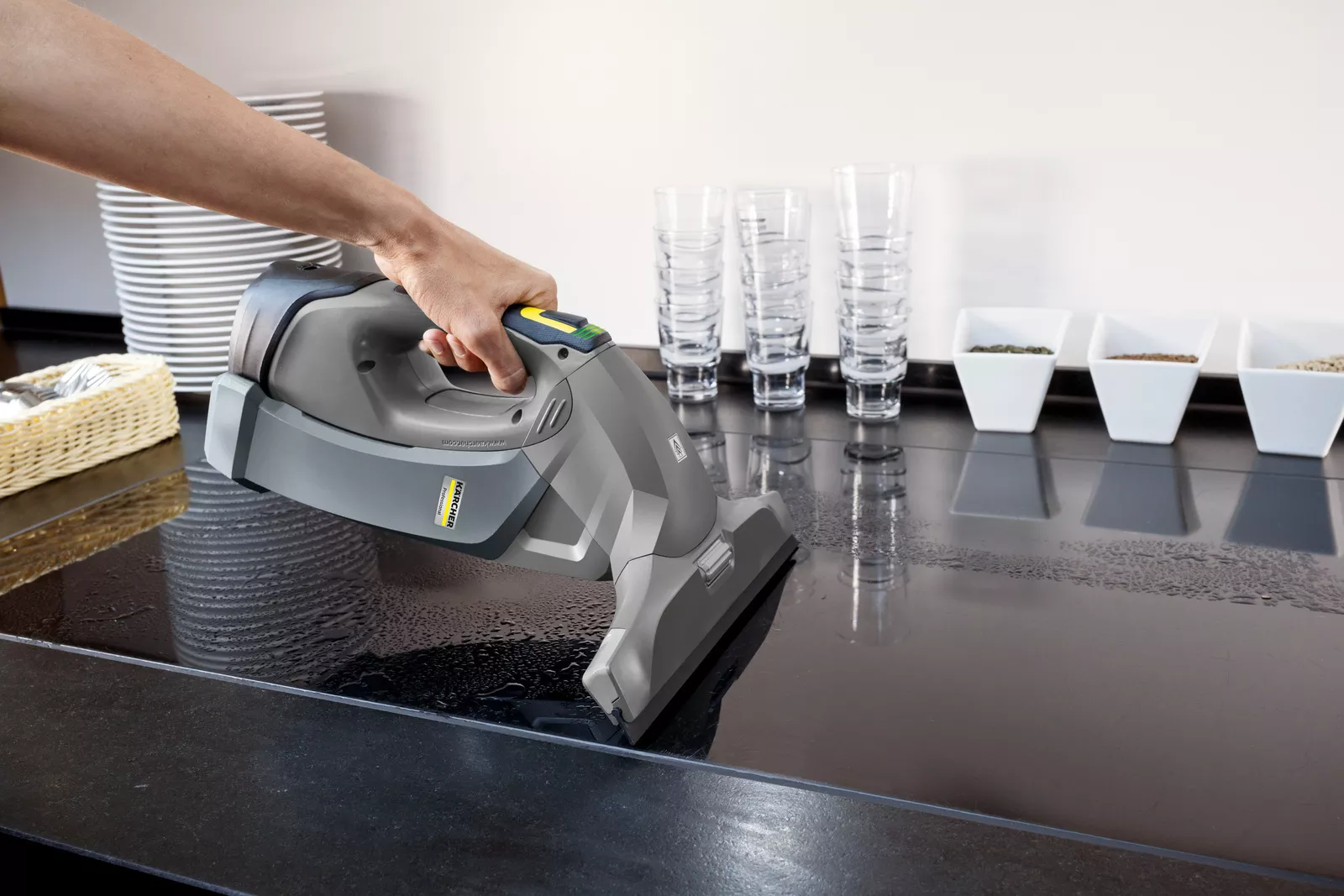 Une main utilise un aspirateur portatif gris pour nettoyer un comptoir noir mouillé. Des assiettes empilées sont visibles.