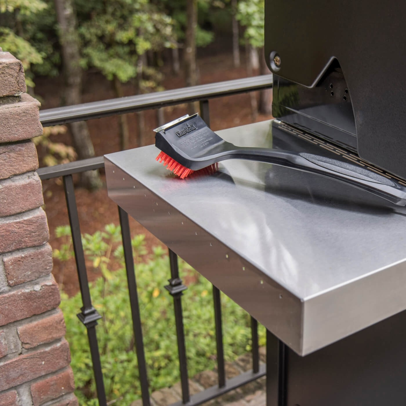 Grillbürste mit roten Borsten auf einem Edelstahltisch neben einem Grill.