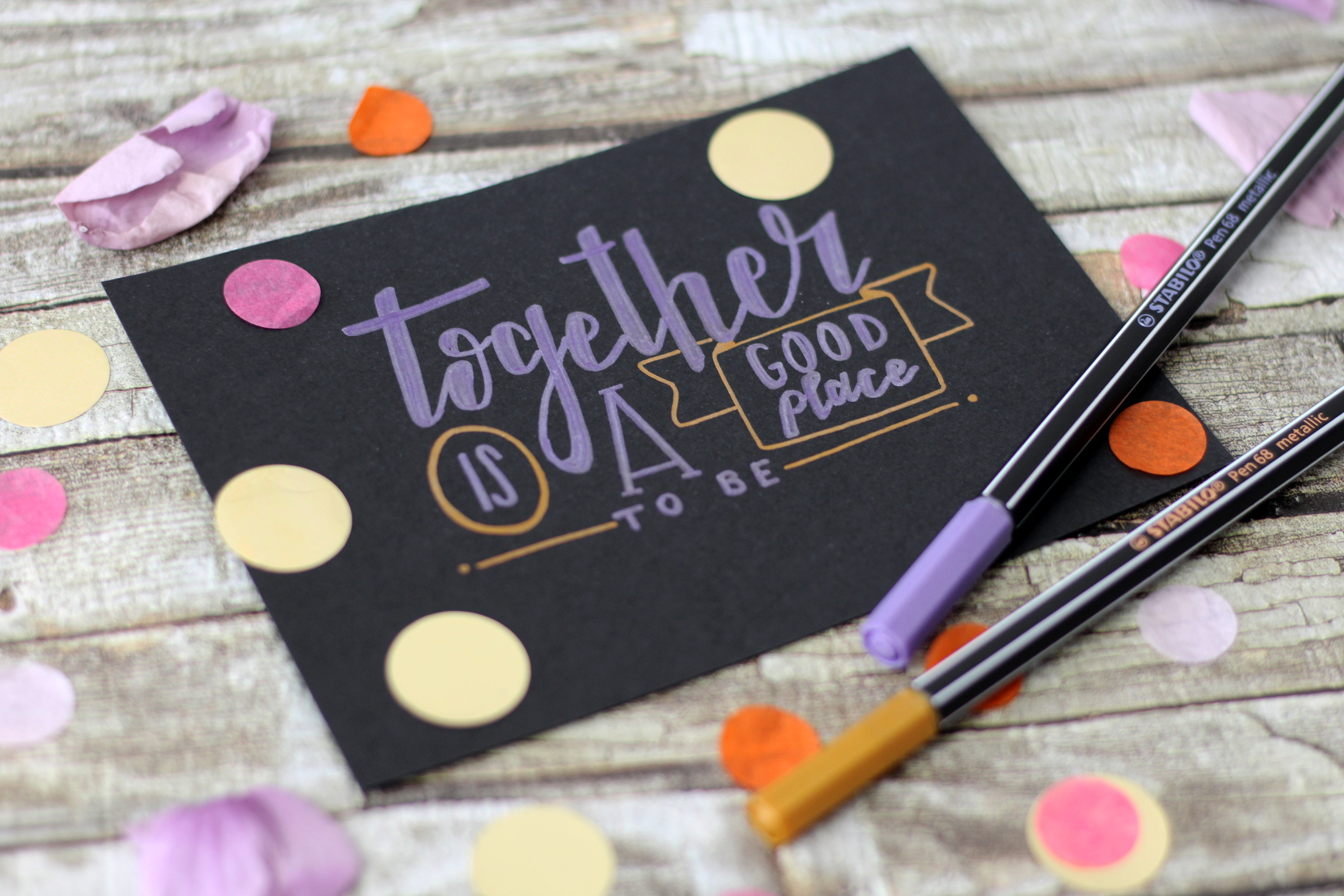 Biglietto nero con testo viola "Together is a good place", coriandoli e penne.
