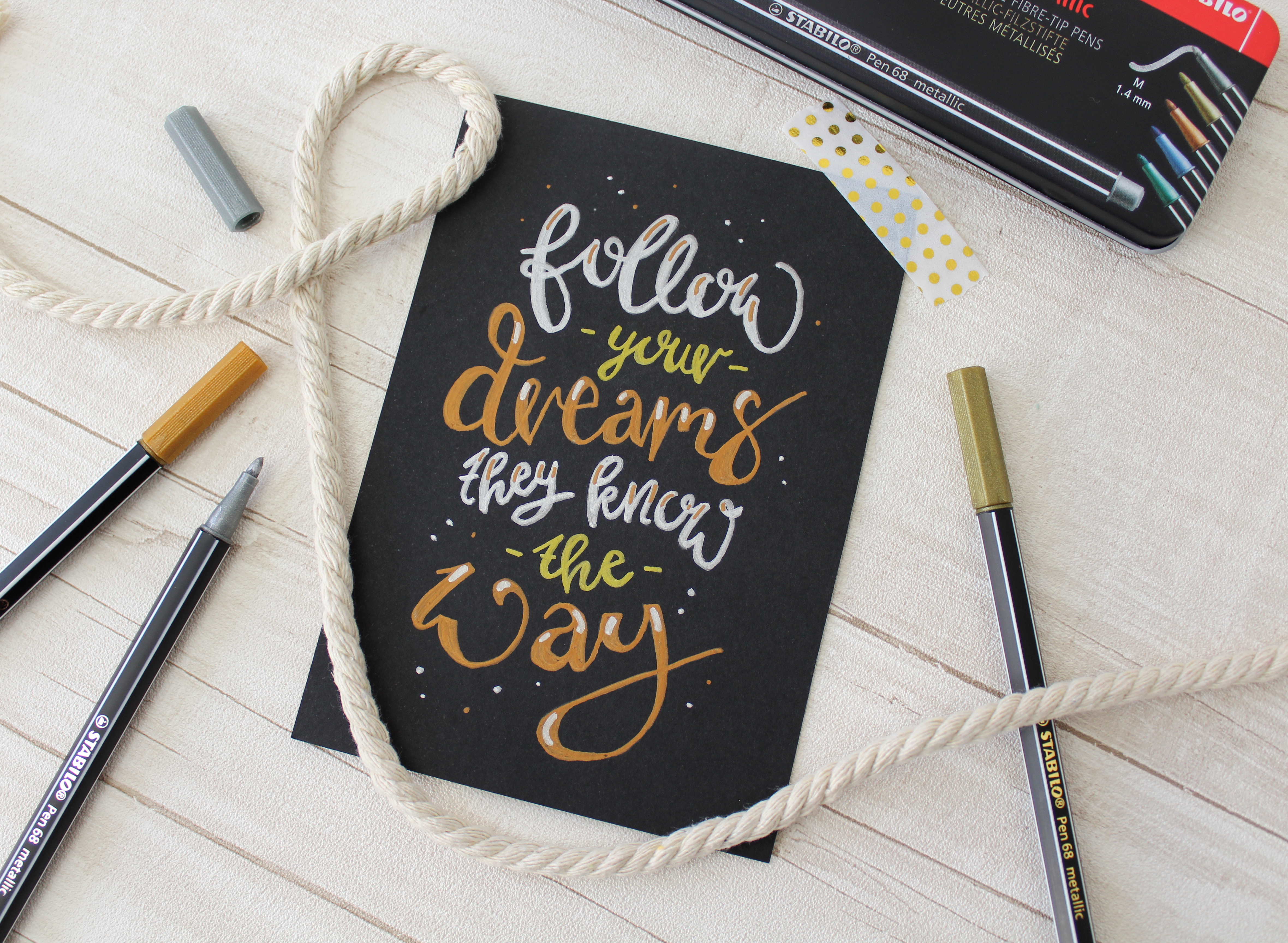 Cartoncino nero con scritta “follow your dreams”, penne e corda su sfondo in legno.