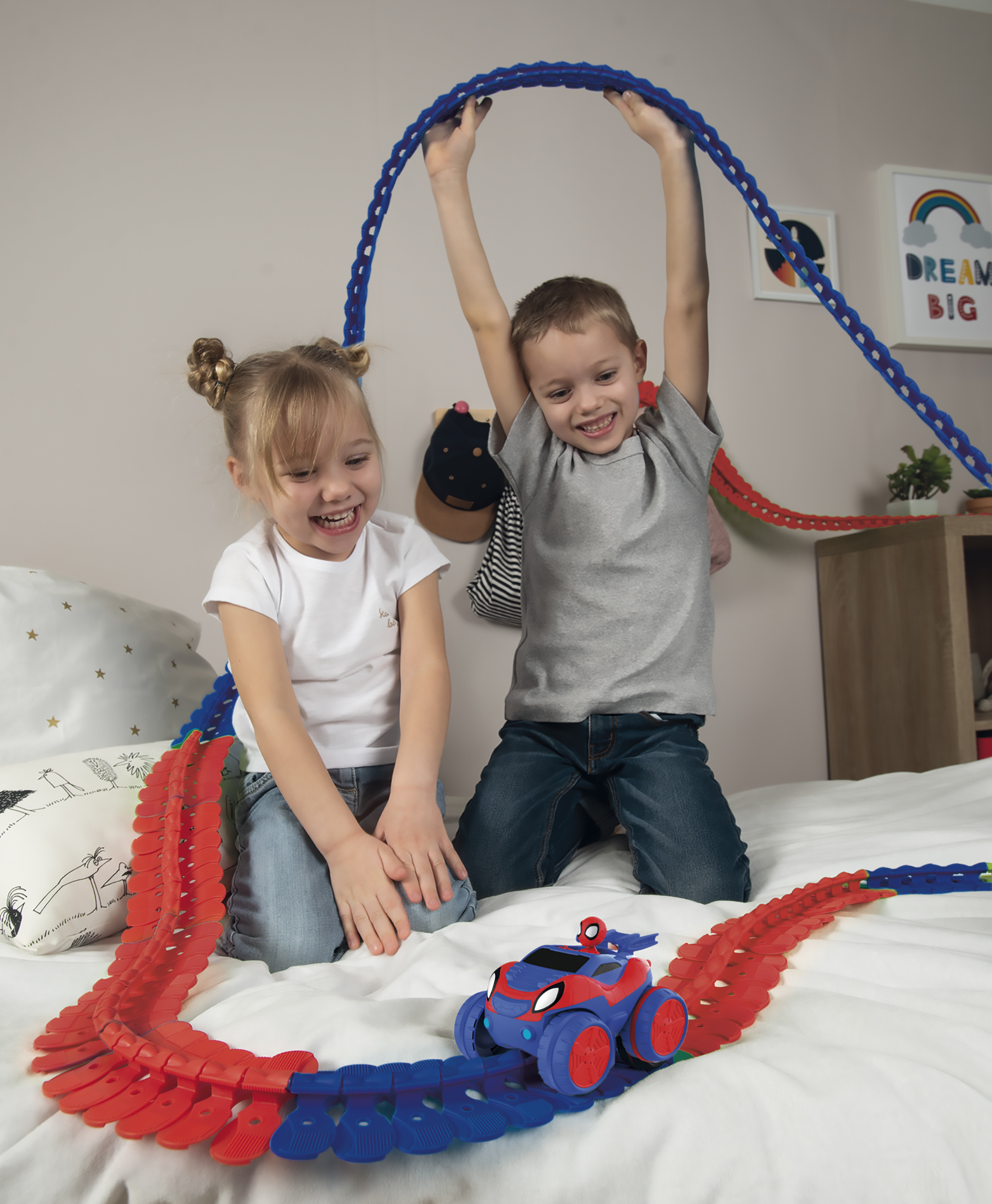 Due bambini giocano con una pista per macchinine su un letto. Un ragazzo tiene in alto una pista blu.