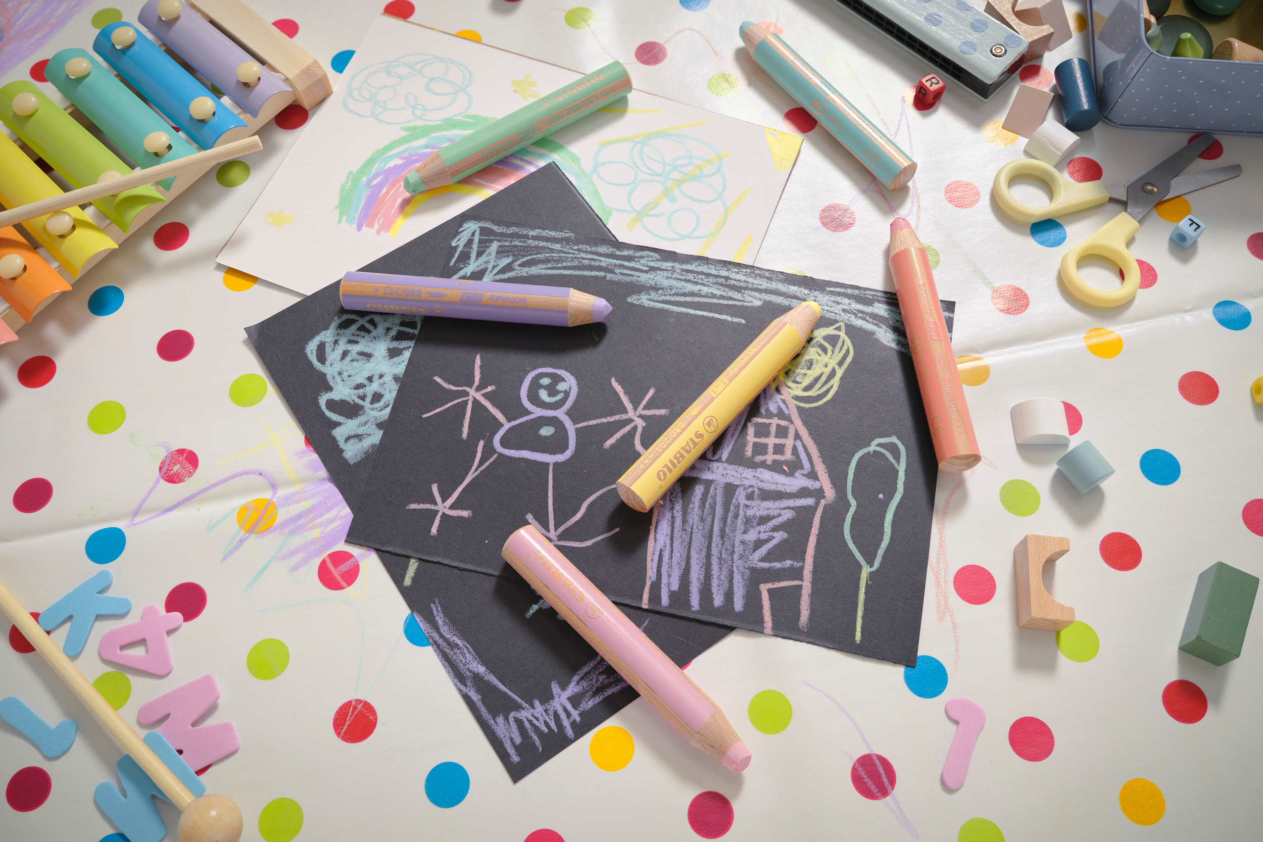 Colorati disegni per bambini con pastelli, xilofono e giocattoli in legno su tavolo a pois.