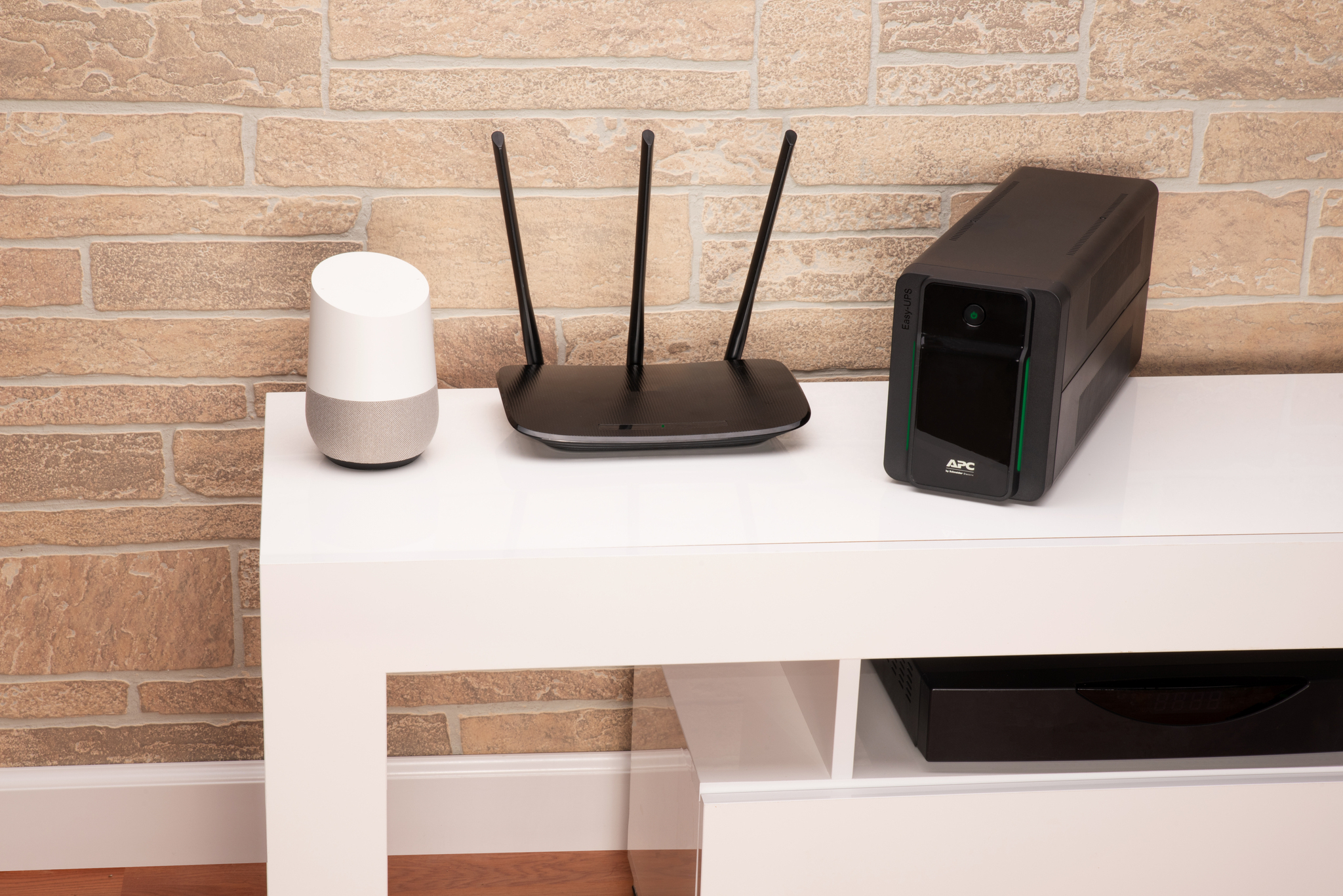 Un Google Home, router e UPS su uno scaffale bianco contro un muro di mattoni.