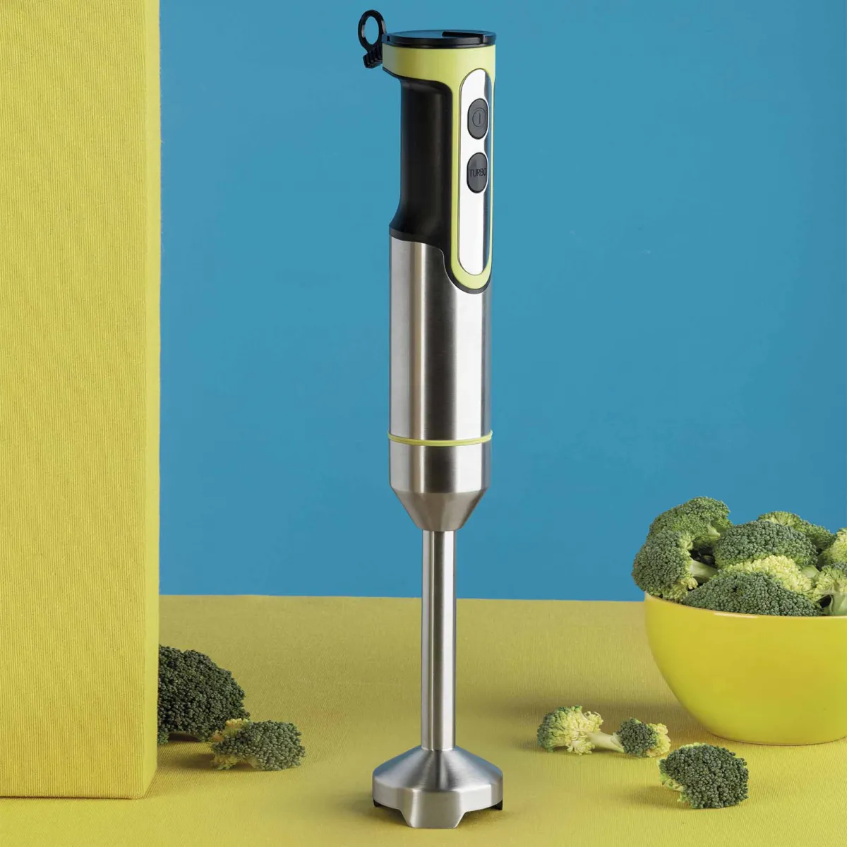 Stalowy blender ręczny z zielonym uchwytem stoi obok żółtej miski z brokułami.