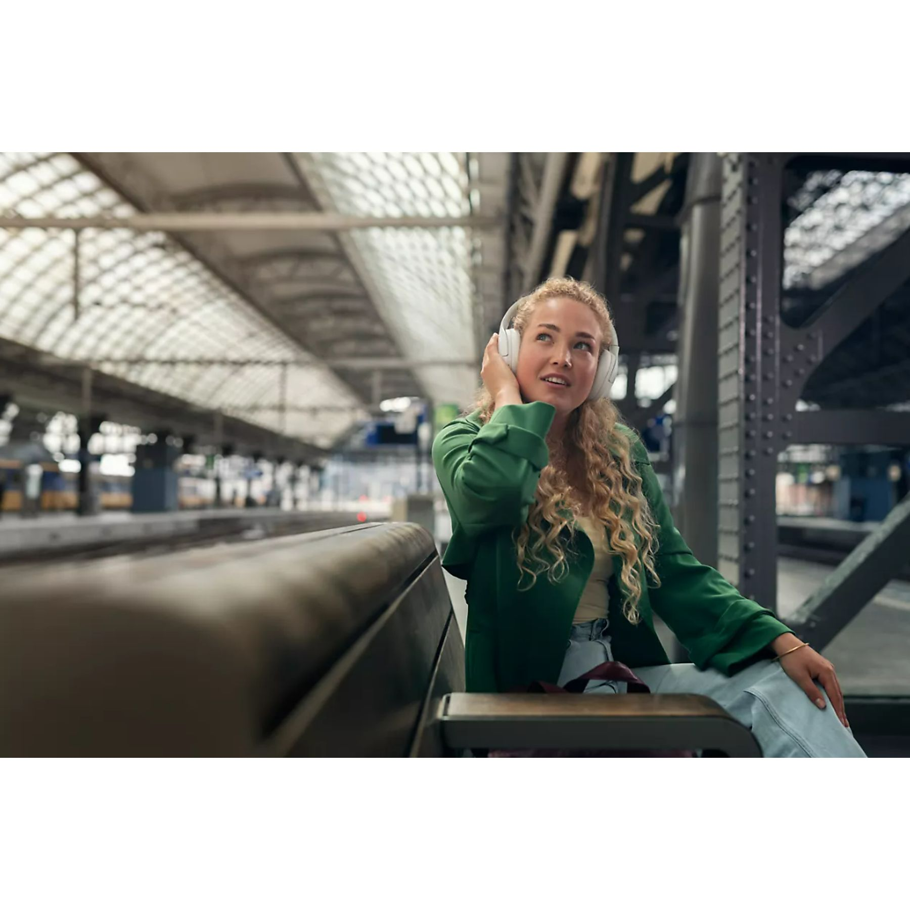 Eine junge Frau mit Kopfhörern sitzt auf einer Bank am Bahnhof und schaut nach oben.