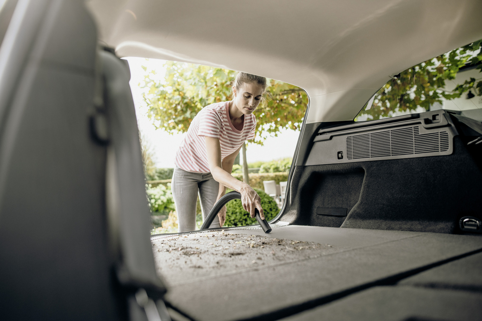 Kobieta odkurza bagażnik samochodu, na zewnątrz drzewa.