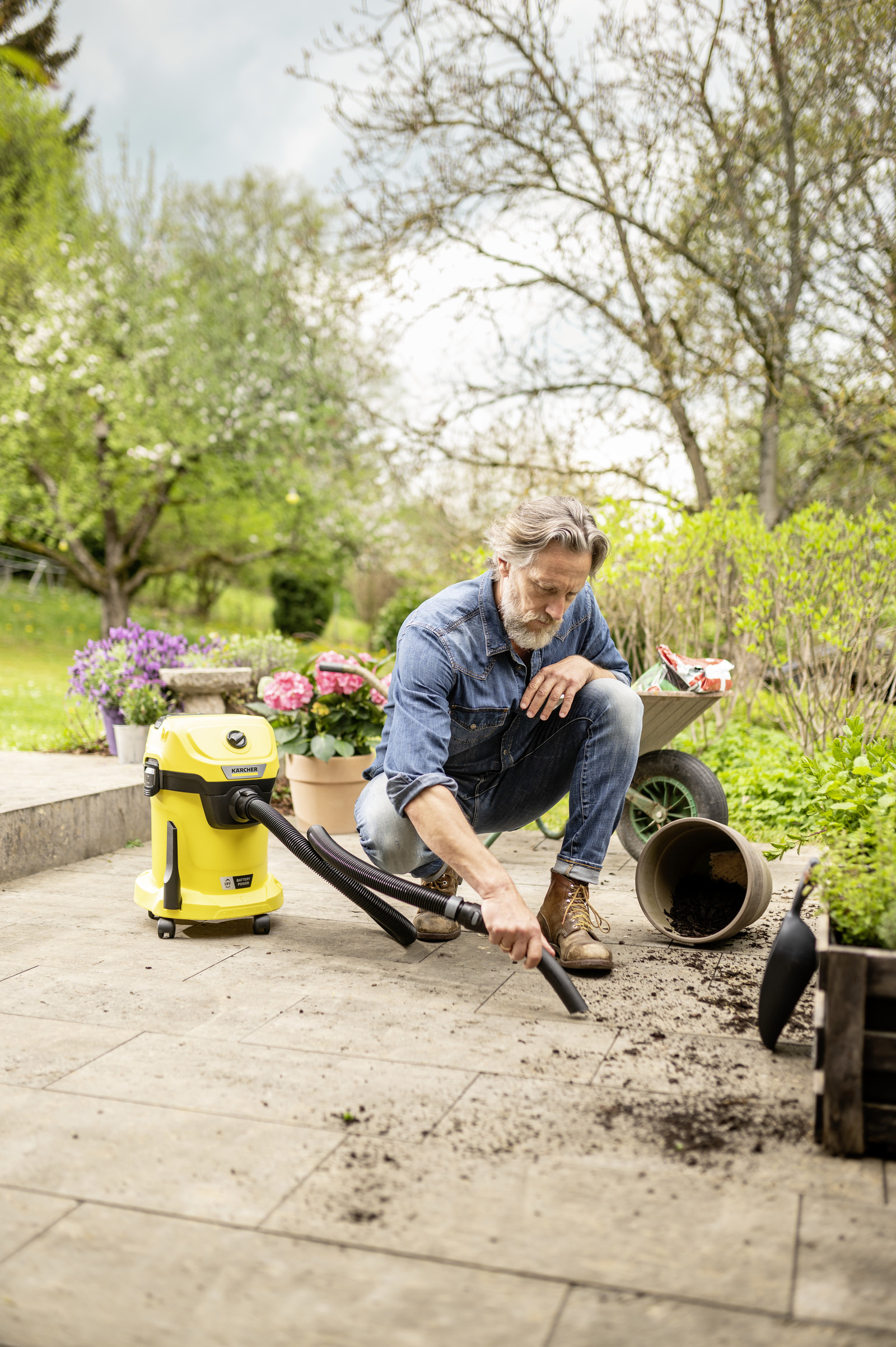 Mann reinigt Hof mit gelbem Kärcher Sauger. Garten mit Blumen und Karre im Hintergrund.