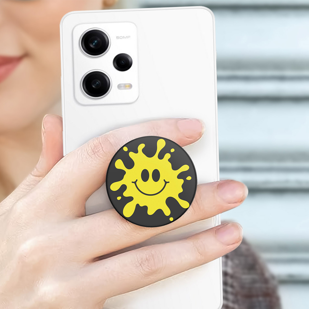 Una mano tiene uno smartphone con un pop socket giallo a faccina sorridente.