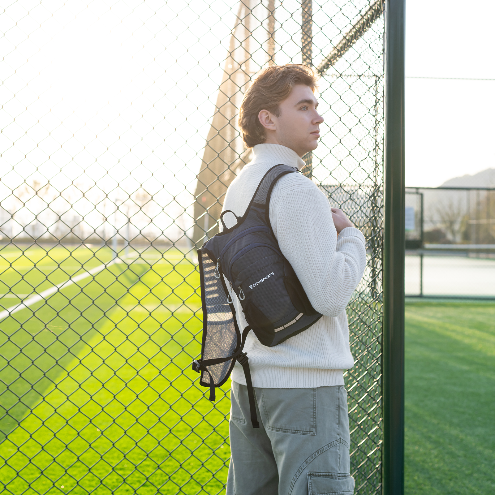 Mężczyzna z plecakiem przed boiskiem piłkarskim i płotem.
