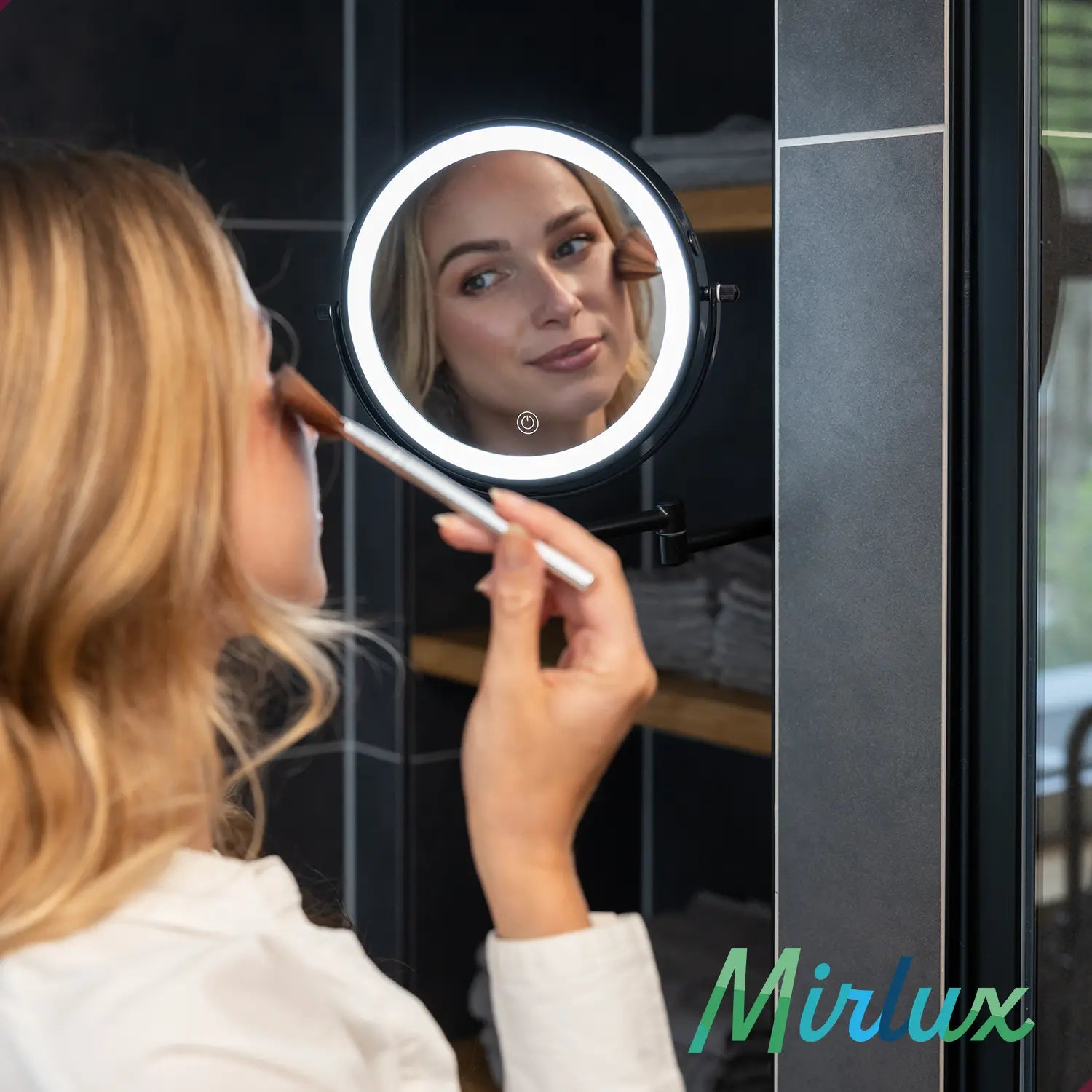 A woman with blonde hair applies makeup in front of a lighted mirror.