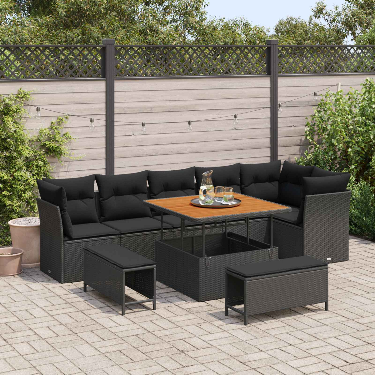 Gartenlounge mit schwarzen Kissen, Holztisch, Bänken auf gepflasterter Terrasse.