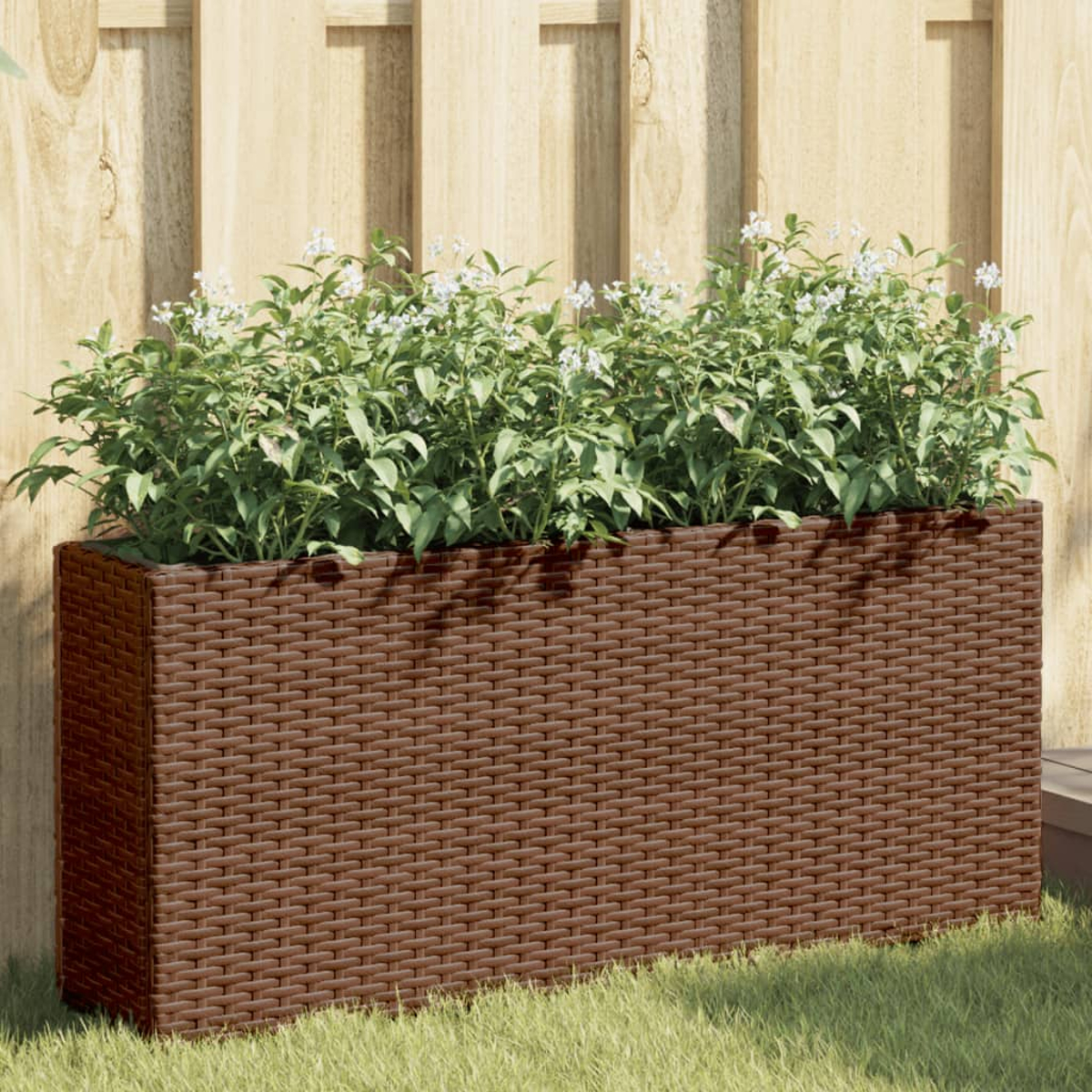 Fioriera in rattan marrone con piante verdi e fiori bianchi davanti a una recinzione in legno.