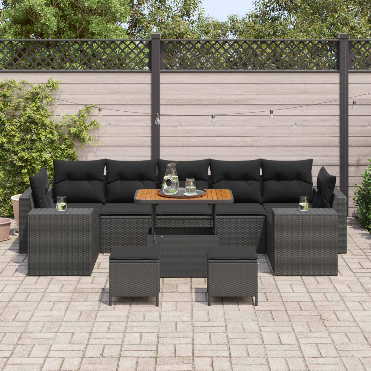 Ein dunkles Rattan-Sofa mit schwarzen Kissen steht auf einer gepflasterten Terrasse.