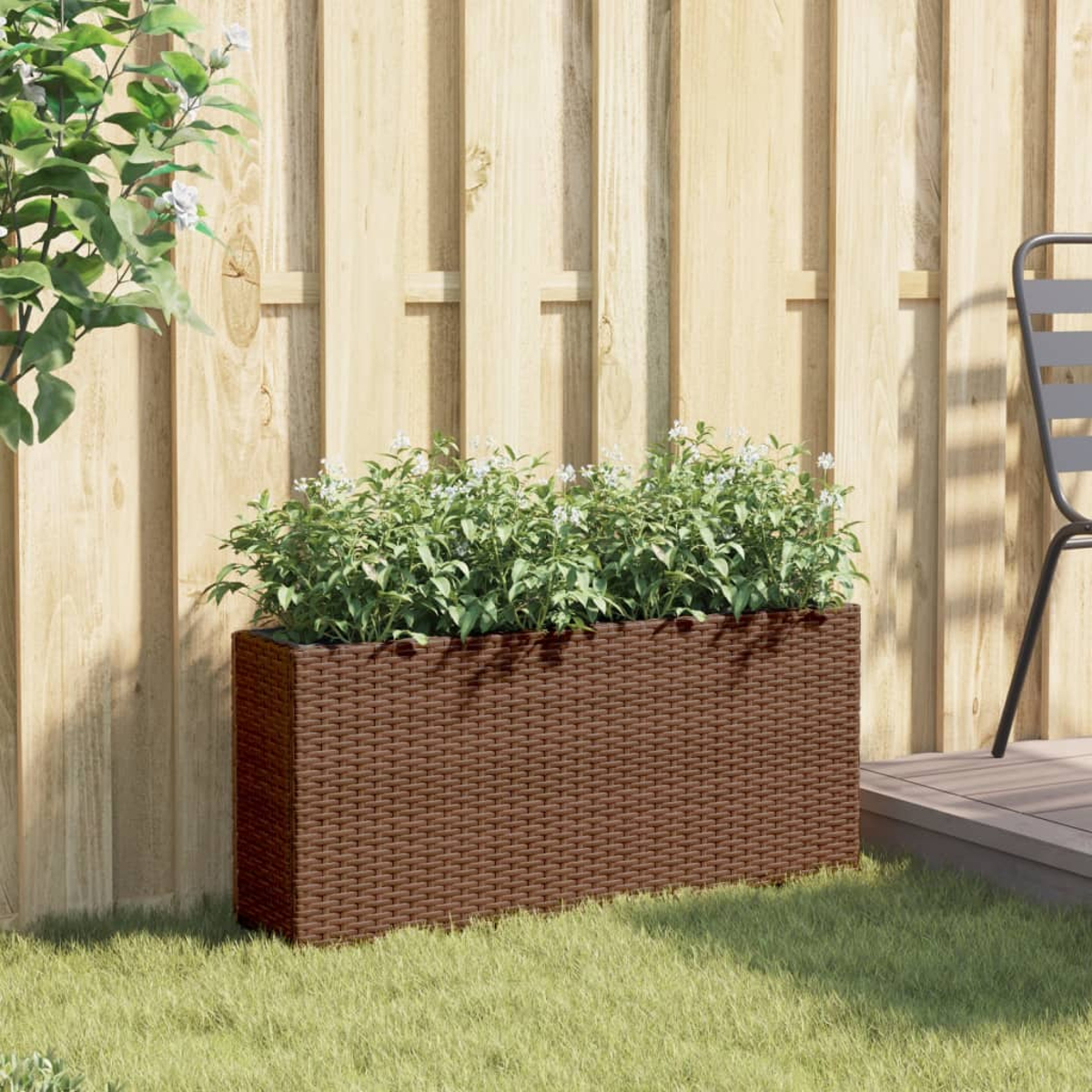 Una fioriera marrone in rattan con piante verdi e fiori bianchi davanti a una recinzione di legno.