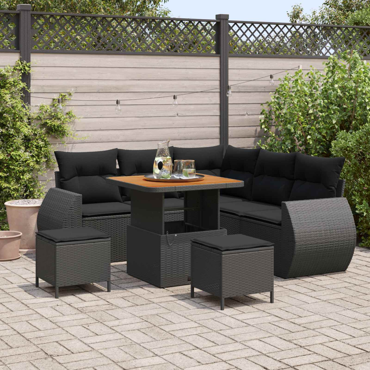 Ein Gartenmöbel-Set mit schwarzem Polster, Tisch und Hockern auf einer Pflasterterrasse.