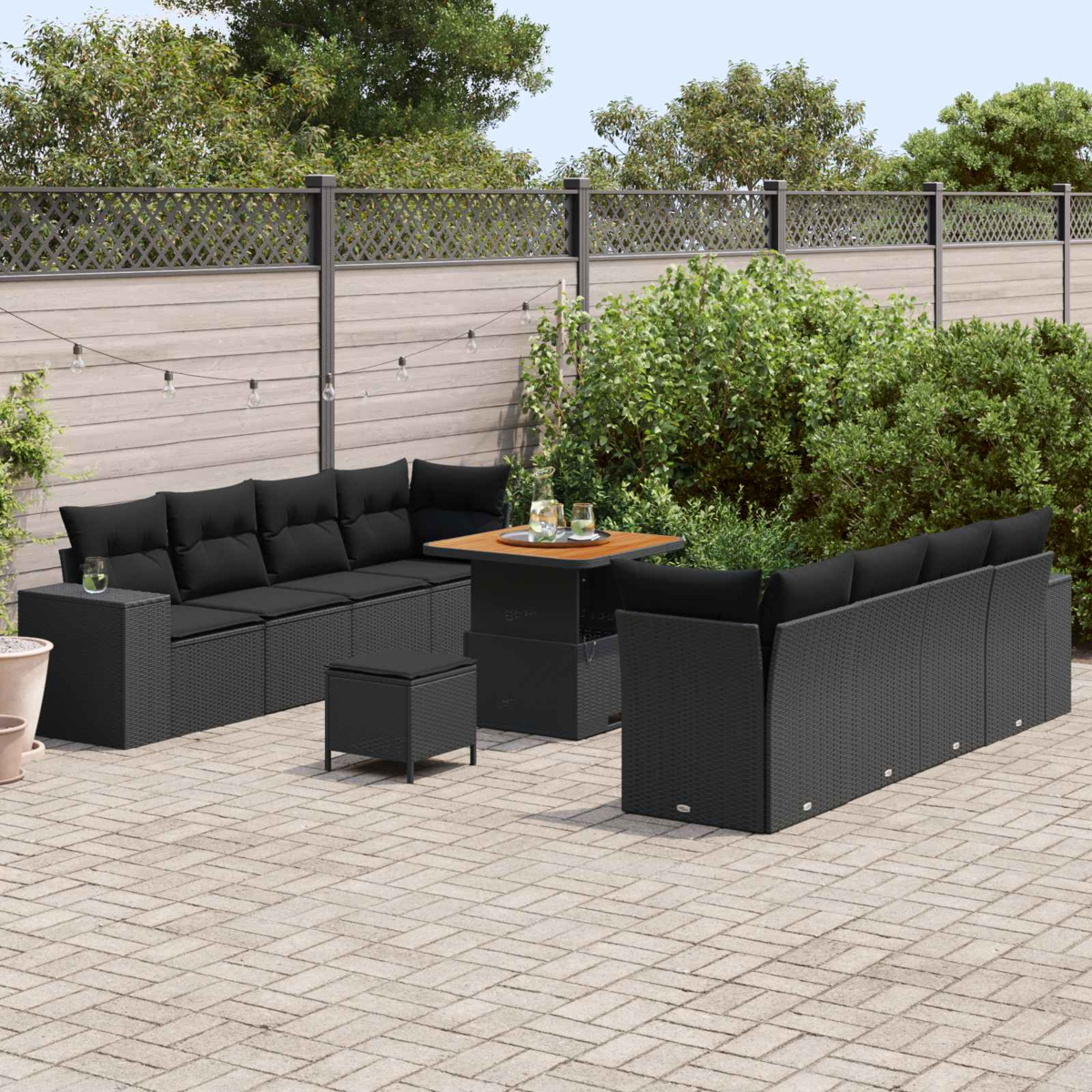 Ein schwarzes Rattan-Sofa und ein Tisch stehen auf einer gepflasterten Terrasse mit Büschen und Zaun.