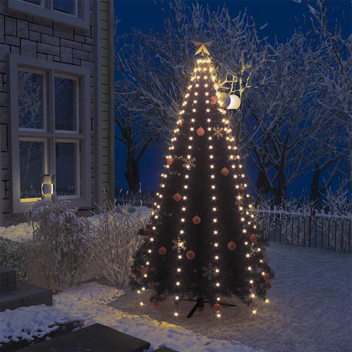 Ein beleuchteter Weihnachtsbaum im Schnee vor einem Haus, nachts.