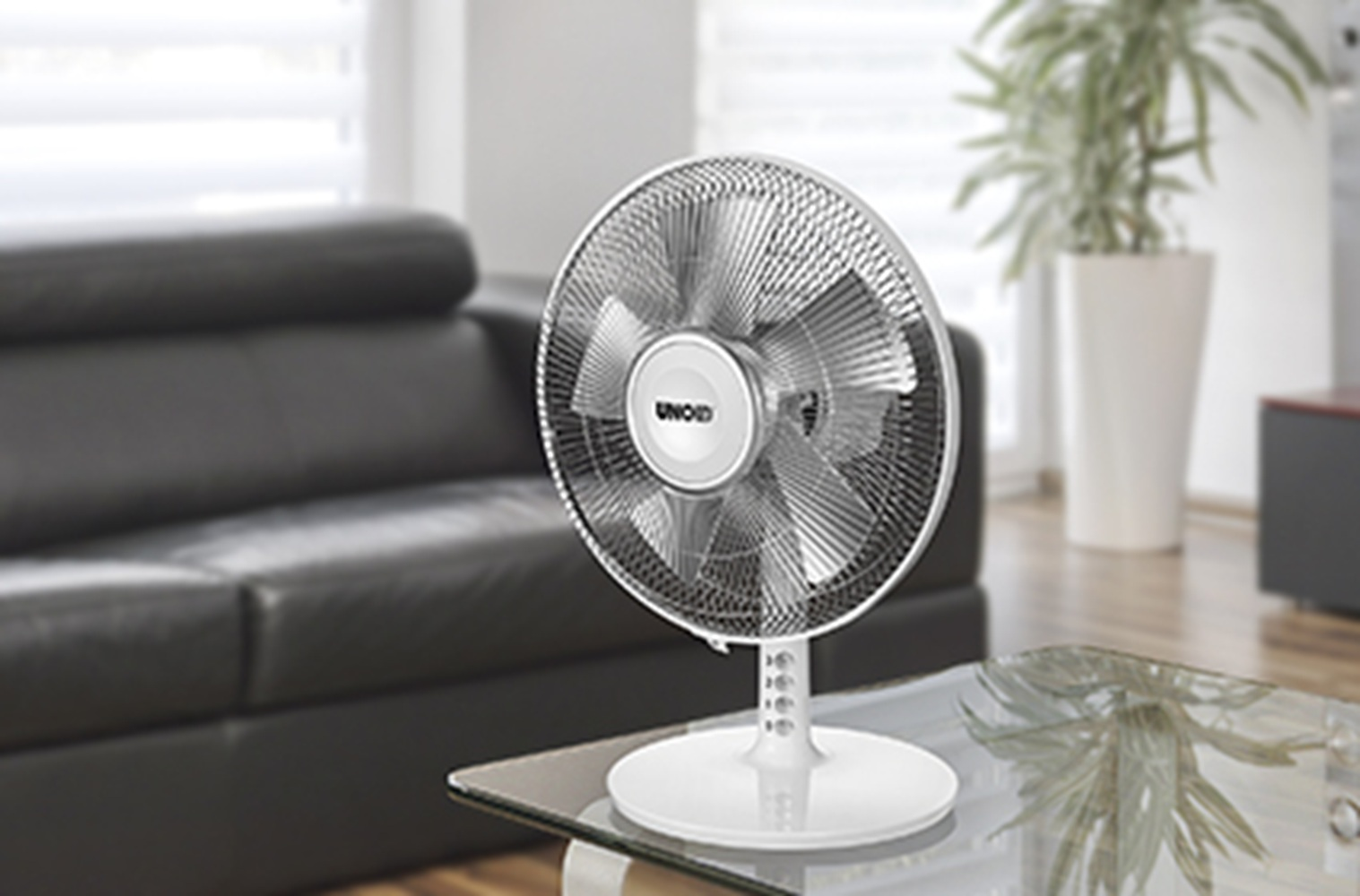 Ein weißer Standventilator steht auf einem Glastisch vor einem schwarzen Sofa.