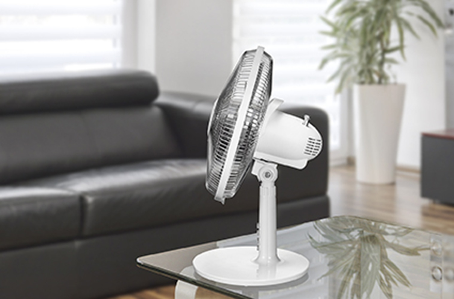 Ein weißer Ventilator steht auf einem Glastisch vor einem schwarzen Sofa.