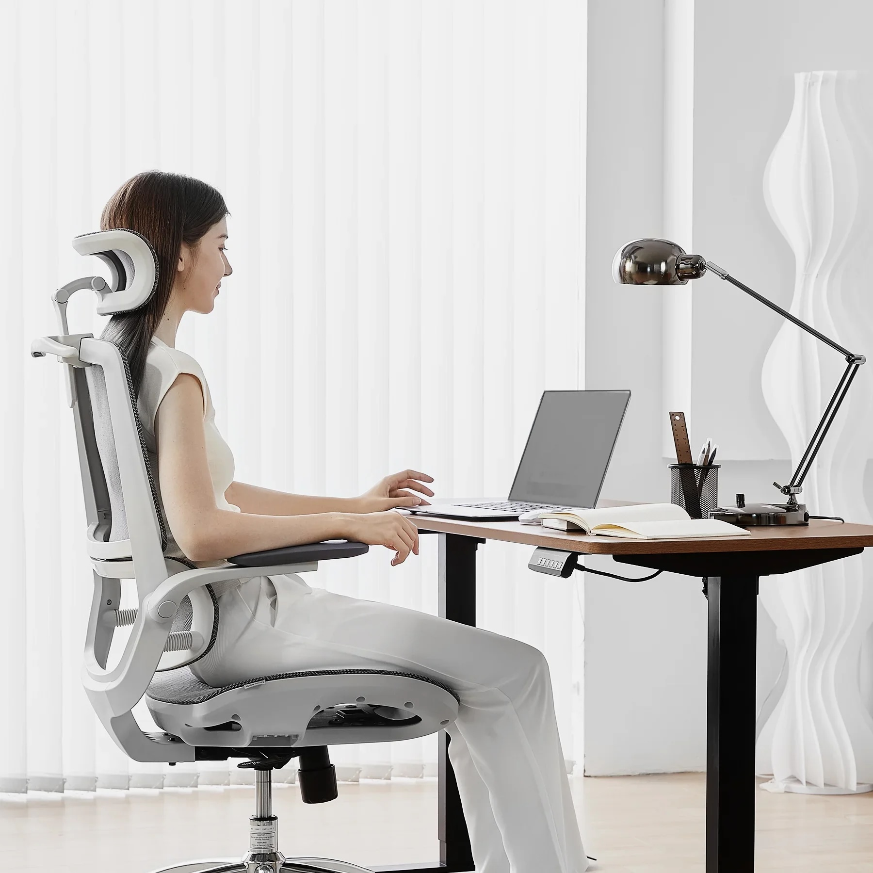 Frau sitzt am Schreibtisch, arbeitet am Laptop, mit Lampe und Notizbuch.