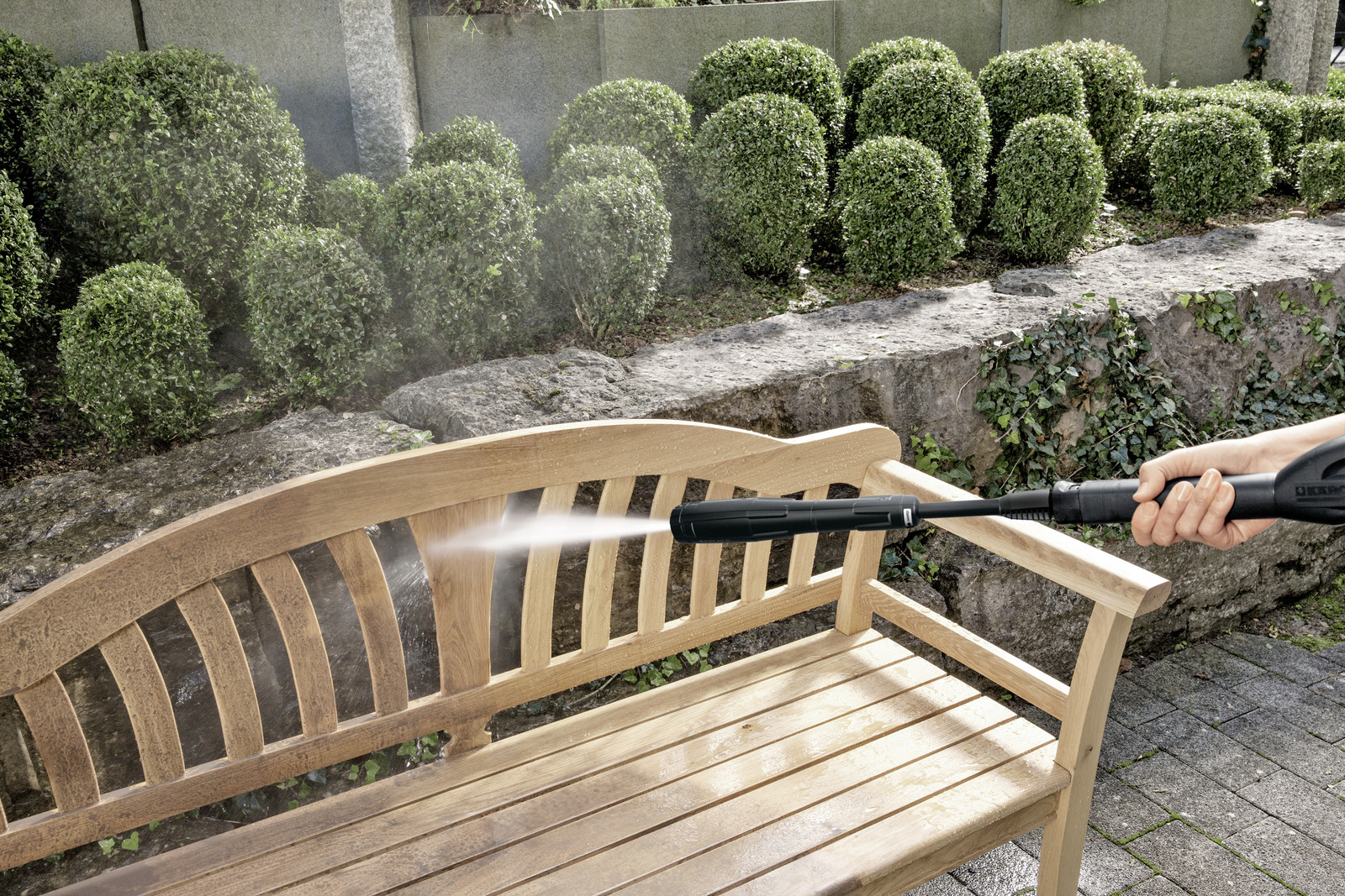 Myjka ciśnieniowa spryskuje wodą drewnianą ławkę w ogrodzie z krzakami w tle.