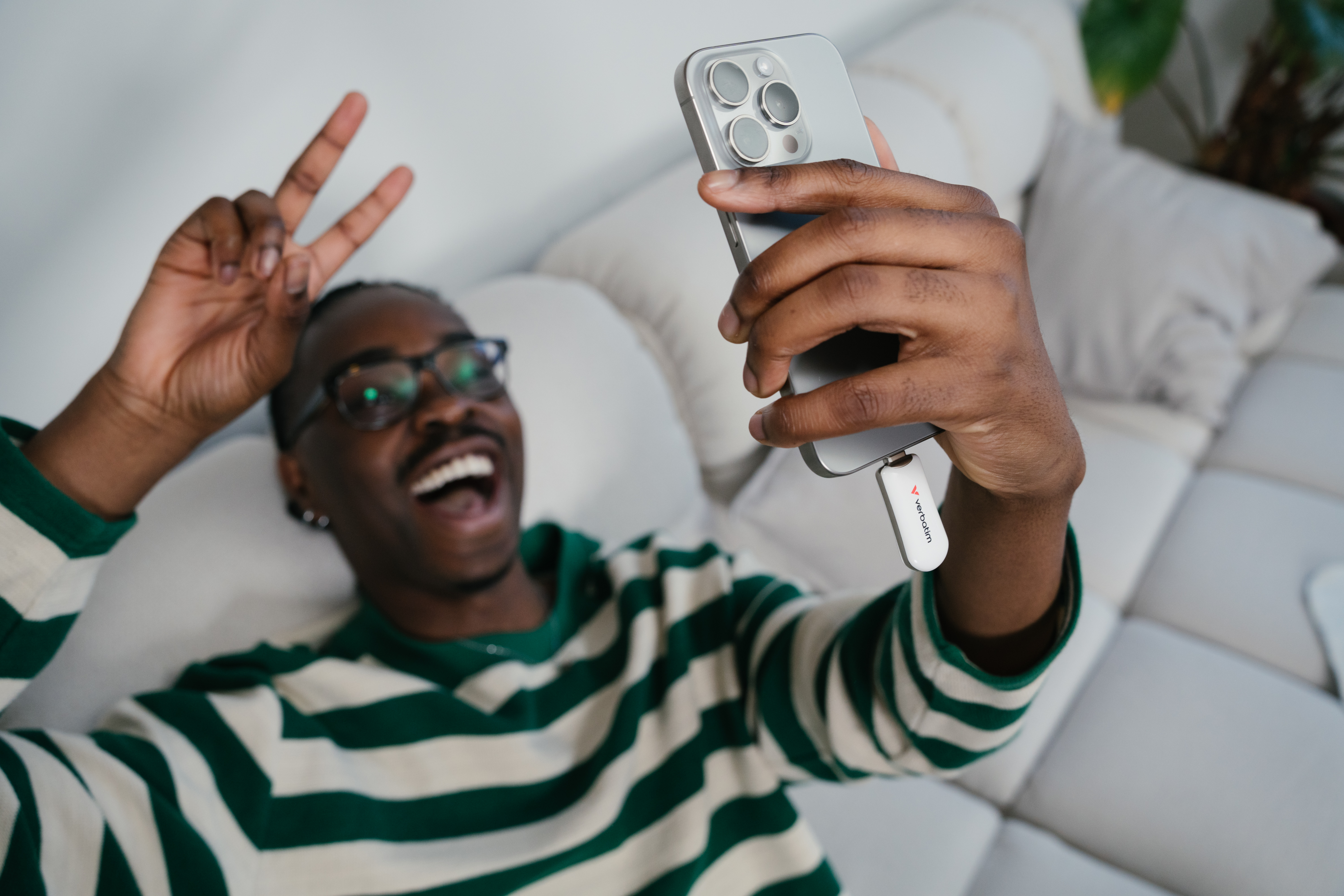 Mężczyzna w okularach na sofie robi sobie selfie telefonem, pokazując dwa palce.
