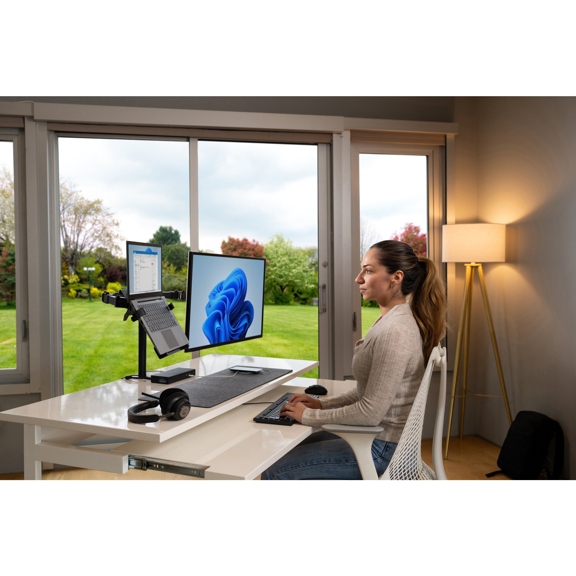 Vrouw aan bureau met dubbele monitor en laptop, uitkijkend op een tuin.