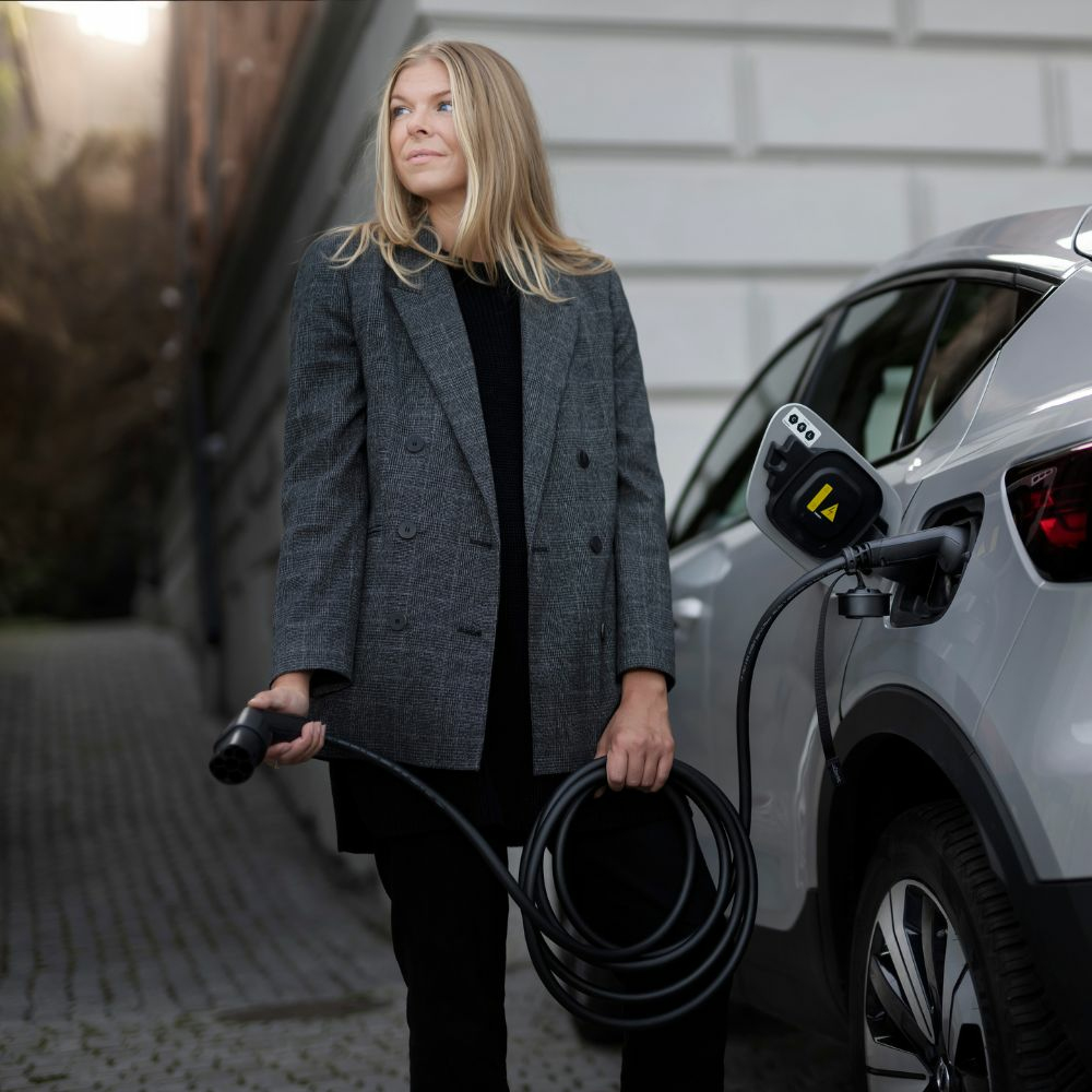 Frau mit Ladekabel steht neben einem Elektroauto, das aufgeladen wird.