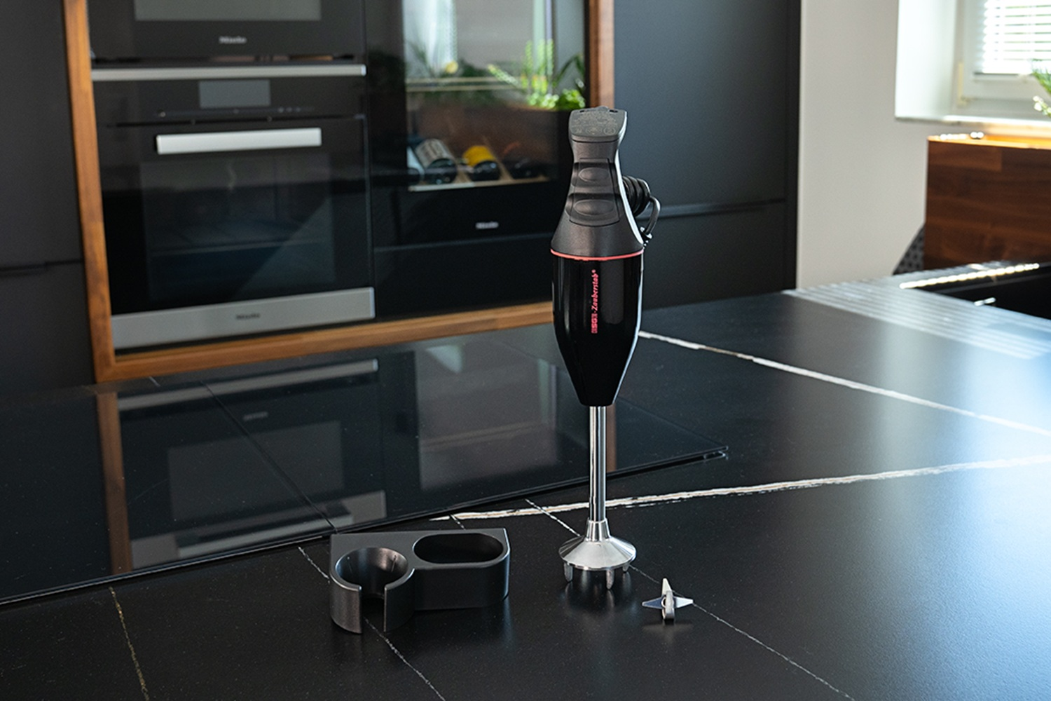 Zwarte staafmixer met rode accenten op een marmeren aanrechtblad, met accessoires ernaast.