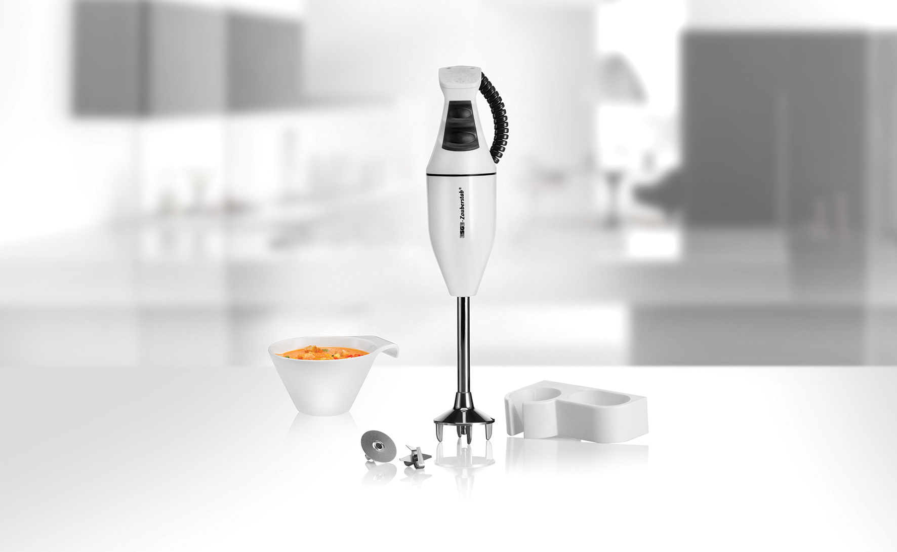 Un frullatore a immersione bianco con accessori, accanto a una ciotola di zuppa arancione.