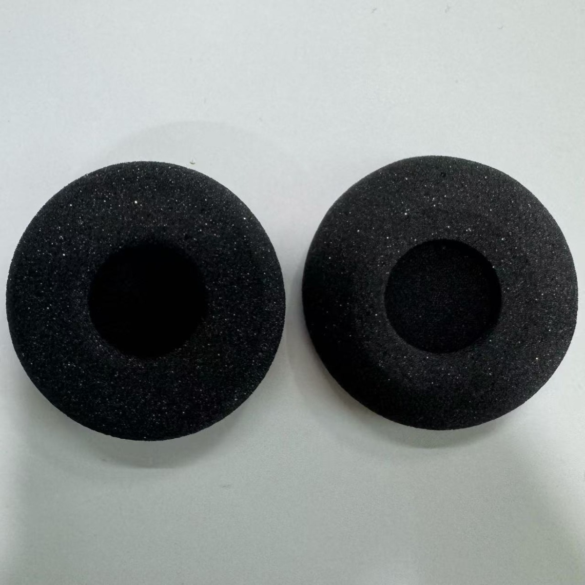 Dos almohadillas negras de espuma para auriculares se muestran una al lado de la otra sobre una superficie blanca.