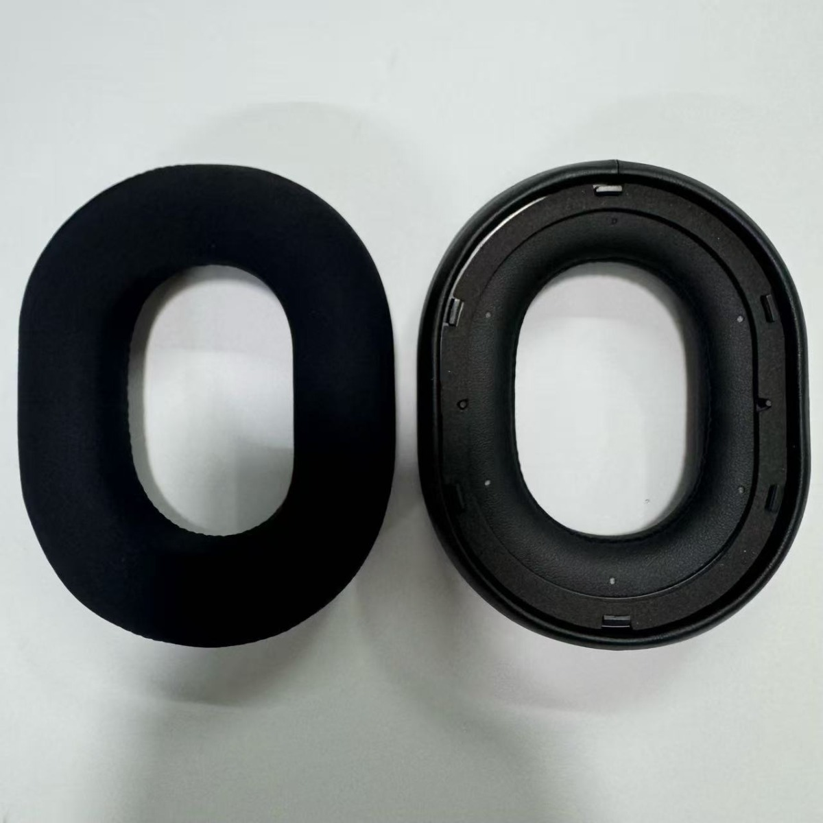 Un cojín de oído negro junto a un marco de repuesto negro para auriculares.