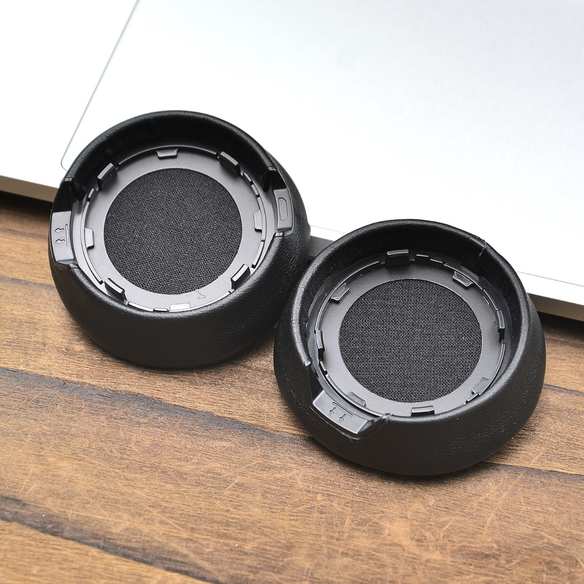 Dos almohadillas negras para auriculares yacen sobre una mesa de madera frente a un portátil.