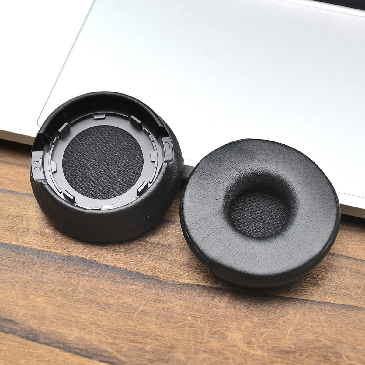 Dos almohadillas negras para auriculares descansan sobre una mesa de madera, parcialmente sobre un portátil.