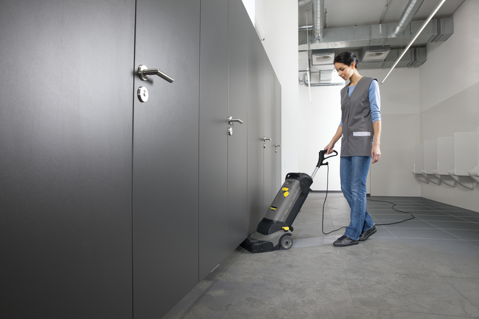 Een vrouw maakt schoon met een schrobzuigmachine in een toiletruimte met donkere deuren.