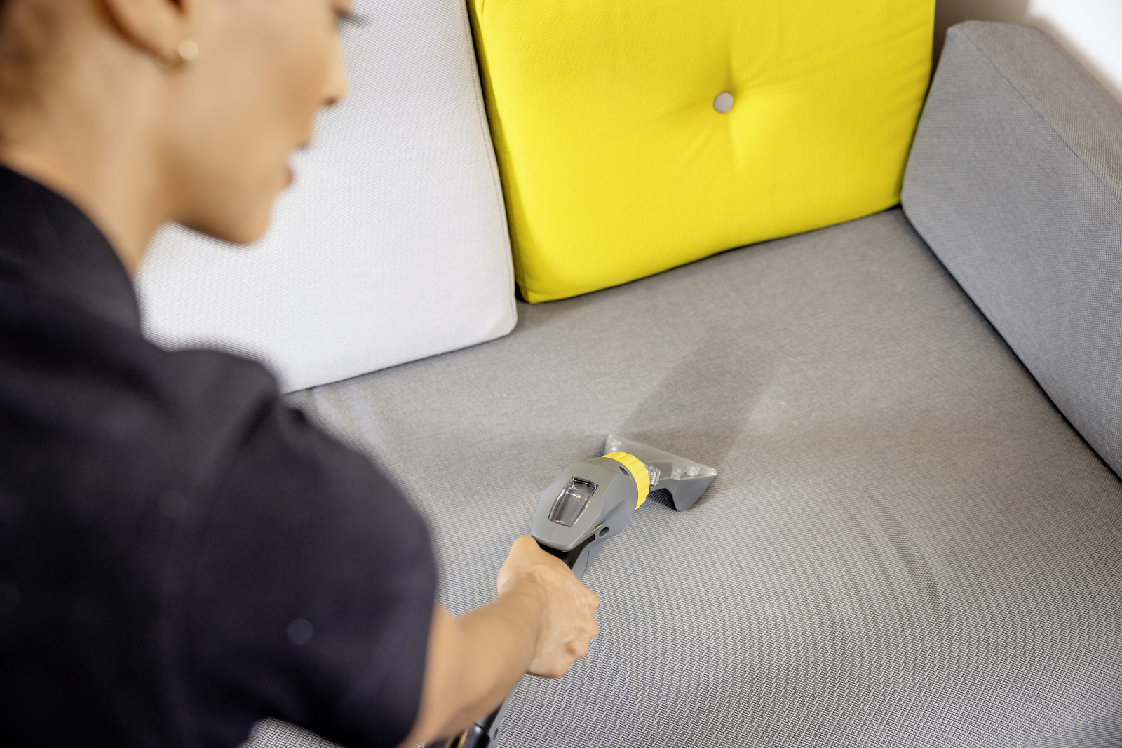 Woman cleaning a grey sofa with a small vacuum cleaner attachment.