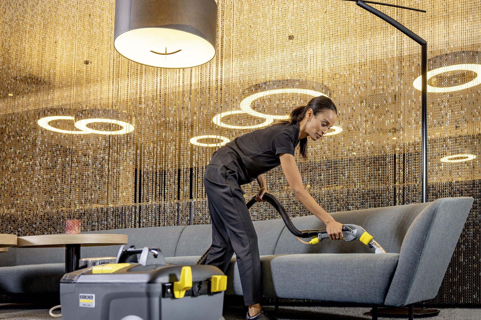 A woman cleans a sofa with a wet/dry vacuum in a modern, lit room.