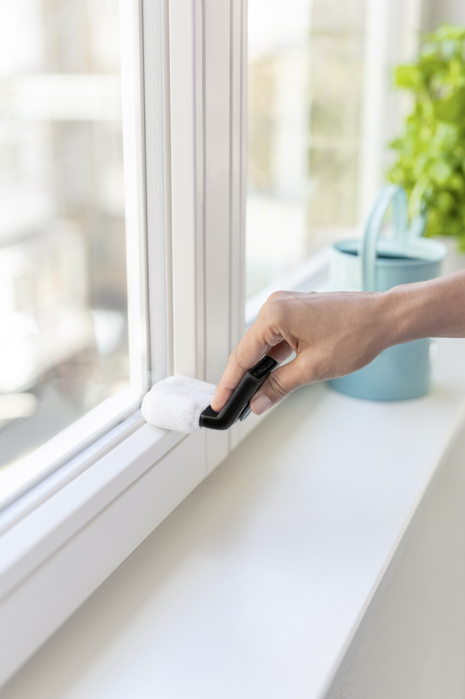 Une main nettoie une fenêtre avec un accessoire blanc, arrosoir bleu en arrière-plan.