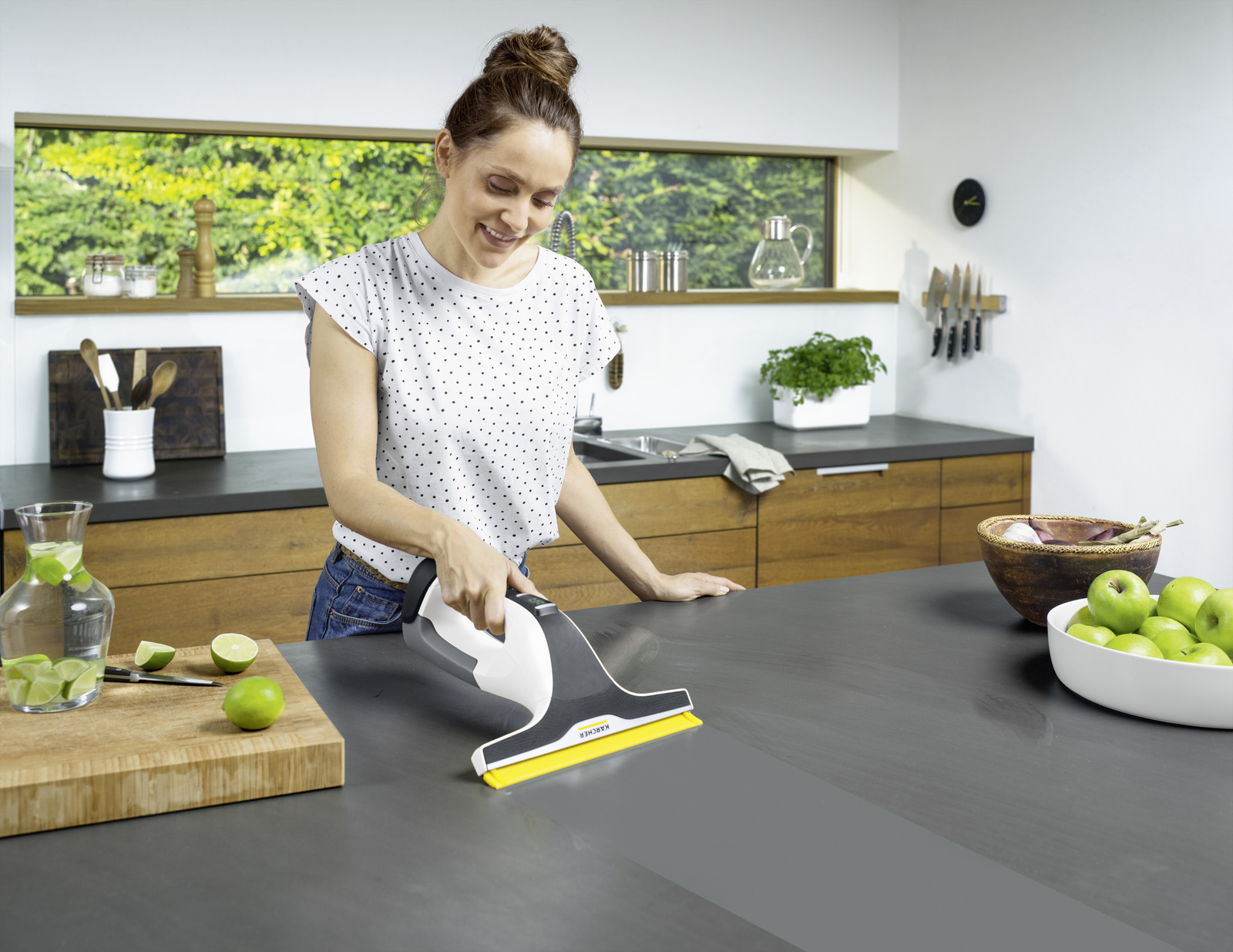 A woman cleans a dark countertop in a modern kitchen with a window cleaner.