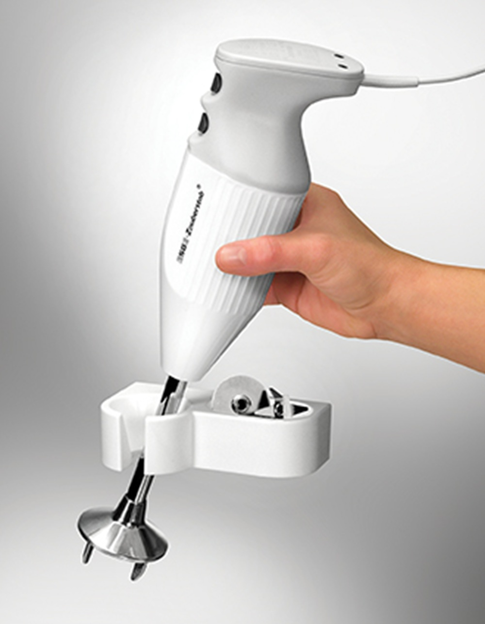 Eine weiße Hand hält einen weißen Stabmixer mit silbernem Aufsatz in einer weißen Halterung.