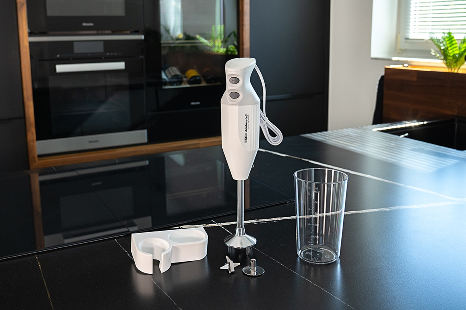 Biały blender ręczny z akcesoriami i szklanką na czarnym blacie.