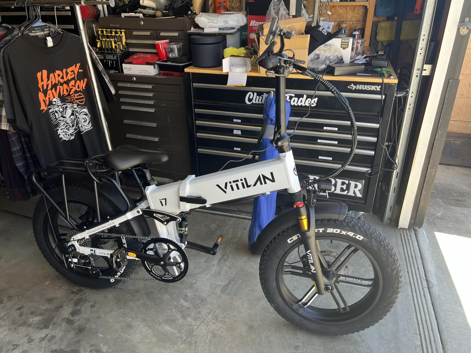 Una e-bike bianca è davanti a cassettiere per attrezzi. Una maglietta Harley-Davidson è appesa nelle vicinanze.