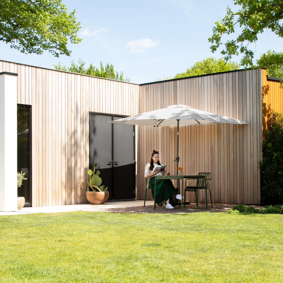 Woman reading a book under a parasol in a garden next to wooden houses.