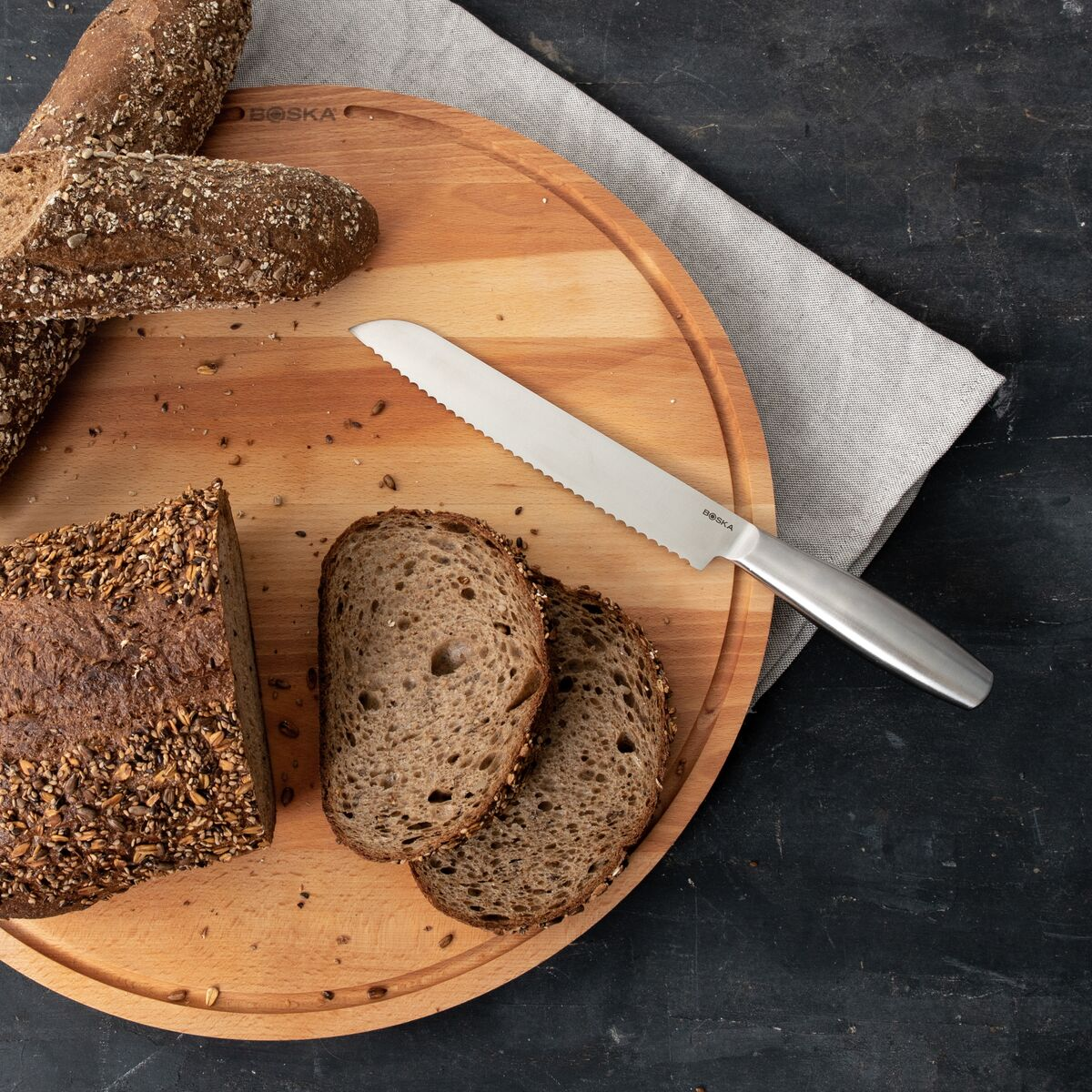 A bread knife rests on a wooden board next to dark bread and slices.