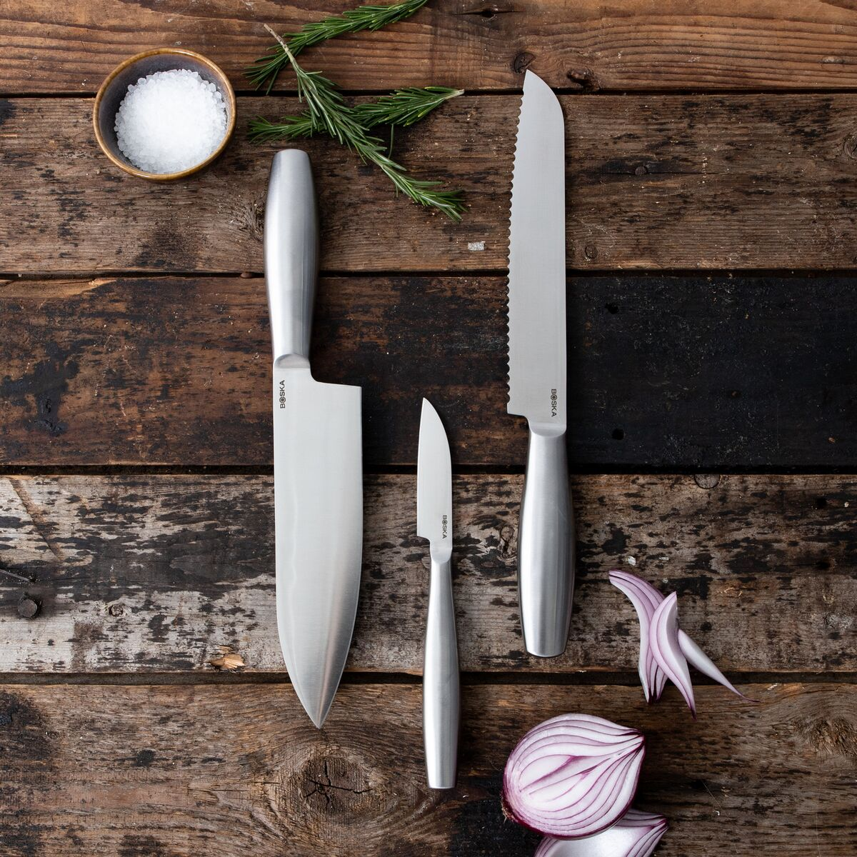 Three kitchen knives, salt, rosemary, and a red onion on wood.