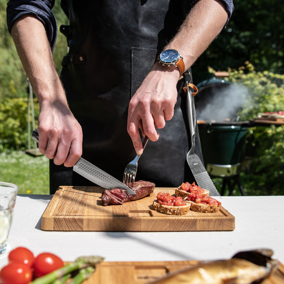 Man cuts steak with knife and fork on wooden board. Grill in background.