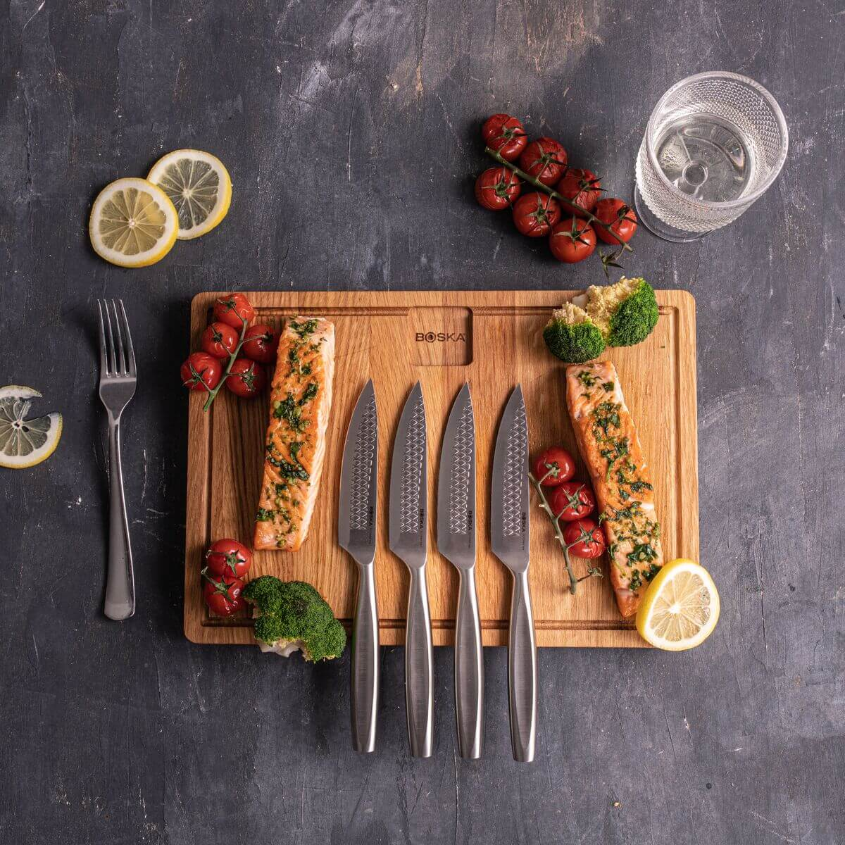 Cooked salmon with tomatoes, broccoli, lemon slices and knives on a wooden board.