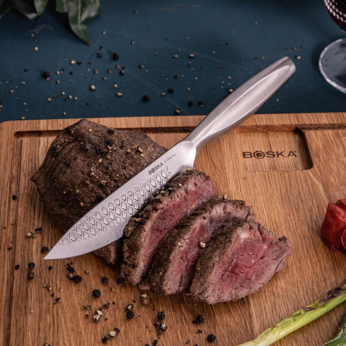 A knife rests on a wooden board next to sliced steak and peppercorns.